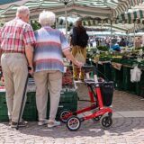 Zwei Senioren stehen mit Gehhilfen und Rollator auf dem Wochenmarkt.