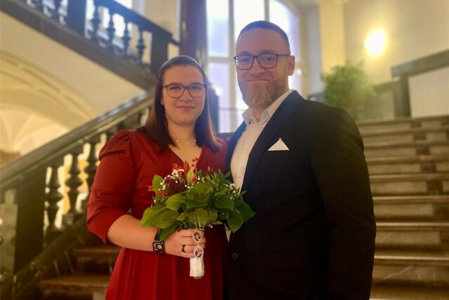 Ein frisch verheiratetes Paar steht auf der Treppe im Dortmunder Standesamt. Beide lächeln in die Kamera, die in rot gekleidete Braut hält einen kleinen Blumenstrauß in der Hand.