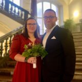 Ein frisch verheiratetes Paar steht  auf der Treppe im Dortmunder Standesamt. Beide lächeln in die Kamera, die in rot gekleidete Braut hält einen kleinen Blumenstrauß in der Hand.
