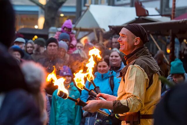 Auf dem Bochumer Weihnachtsmarkt steht ein Mann in mittelalterlicher Kleidung, der drei Fackeln in Händen hält und von zahlreichen Zuschauern umringt ist.