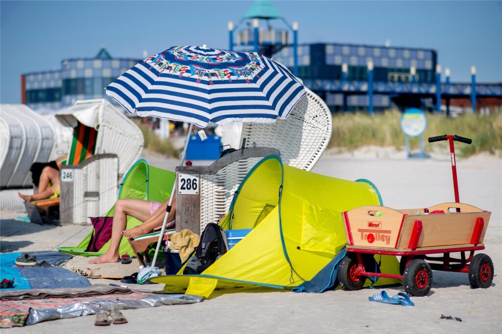 Ein Bollerwagen, ein Strandzelt und ein Sonnenschirm stehen bei sonnigem Wetter neben einem Strandkorb am Strand. Ein Bollerwagen, ein Strandzelt und ein Sonnenschirm stehen bei sonnigem Wetter neben einem Strandkorb am Strand.