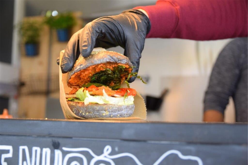 Eine Hand hebt die obere Brötchenhälfte eines Burgers hoch. Belegt ist er mit verschiedenem Gemüse und Soßen.