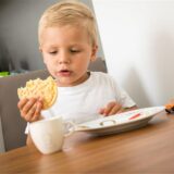 Ein Kind sitzt am Tisch und hält eine Reiswaffel in der Hand und schaut sie sich an.