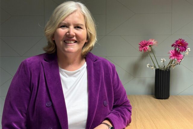 Antje Joest steht in violettem Mantel vor einem neutralen Hintergrund, neben ihr eine Blumenvase