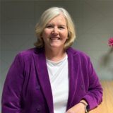 Antje Joest steht in violettem Mantel vor einem neutralen Hintergrund, neben ihr eine Blumenvase