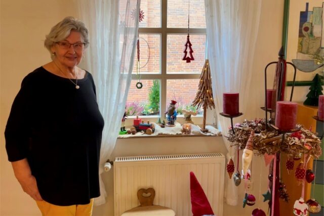 Eine Seniorin steht an einem weihnachtlich dekorierten Fenster.