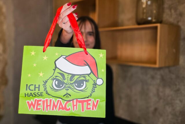 Eine Weihnachtsmarkt-Händlerin hält eine Tüte mit dem Grinch und der Aufschrift „Ich hasse Weihnachten“ in die Höhe.
