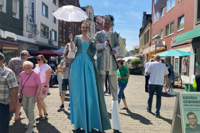 Ein Paar in historischer Kleidung posiert im Freien. Die Frau hält einen weißen Sonnenschirm, während der Mann einen Gehstock trägt.