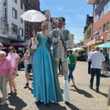 Ein Paar in historischer Kleidung posiert im Freien. Die Frau hält einen weißen Sonnenschirm, während der Mann einen Gehstock trägt.