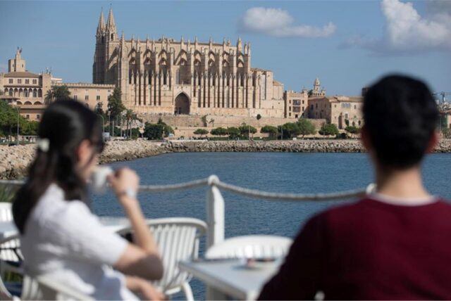 An einem sonnigen Tag in Palma nehmen zwei Personen gegenüber der Kathedrale von Palma einen Drink auf der Terrasse eines Restaurants.