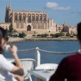 An einem sonnigen Tag in Palma nehmen zwei Personen gegenüber der Kathedrale von Palma einen Drink auf der Terrasse eines Restaurants.