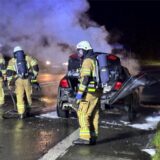 Ein verkohltes Autowrack steht auf einer Straße bei Herten. Drumherum stehen Feuerwehrleute in voller Montur.