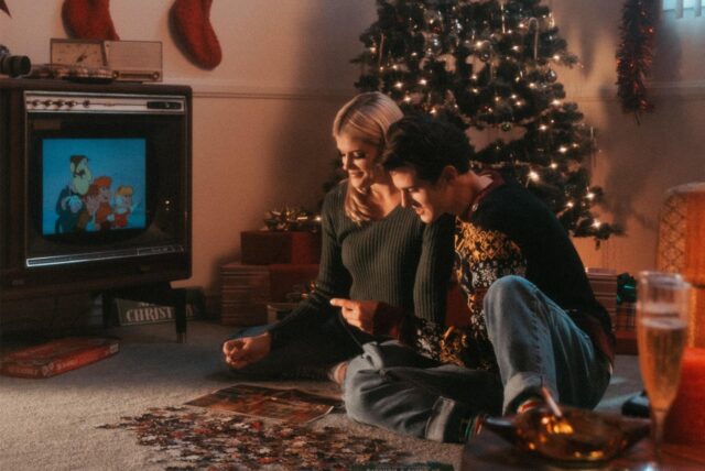 Ein Mann und eine Frau sitzen in einem weihnachtlich geschmückten Wohnzimmer auf dem Boden und gucken einen Weihnachtsfilm.