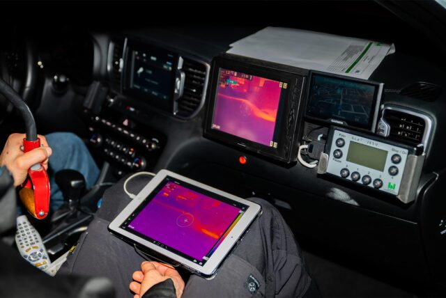 Innenansicht eines Autos, in dem der Beifahrer einen Monitor mit einem Wärmebild auf dem Schoß hält.