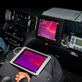 Innenansicht eines Autos, in dem der Beifahrer einen Monitor mit einem Wärmebild auf dem Schoß hält.