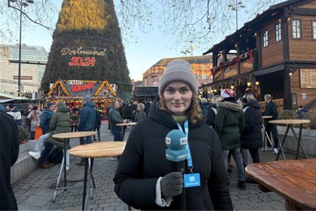 RN-Reporterin Paula Weber steht auf dem Weihnachtsmarkt in Dortmund vor dem großen Weihnachtsbaum, der 2024 für viele verschiedene Meinungen sorgt.