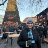 RN-Reporterin Paula Weber steht auf dem Weihnachtsmarkt in Dortmund vor dem großen Weihnachtsbaum, der 2024 für viele verschiedene Meinungen sorgt.
