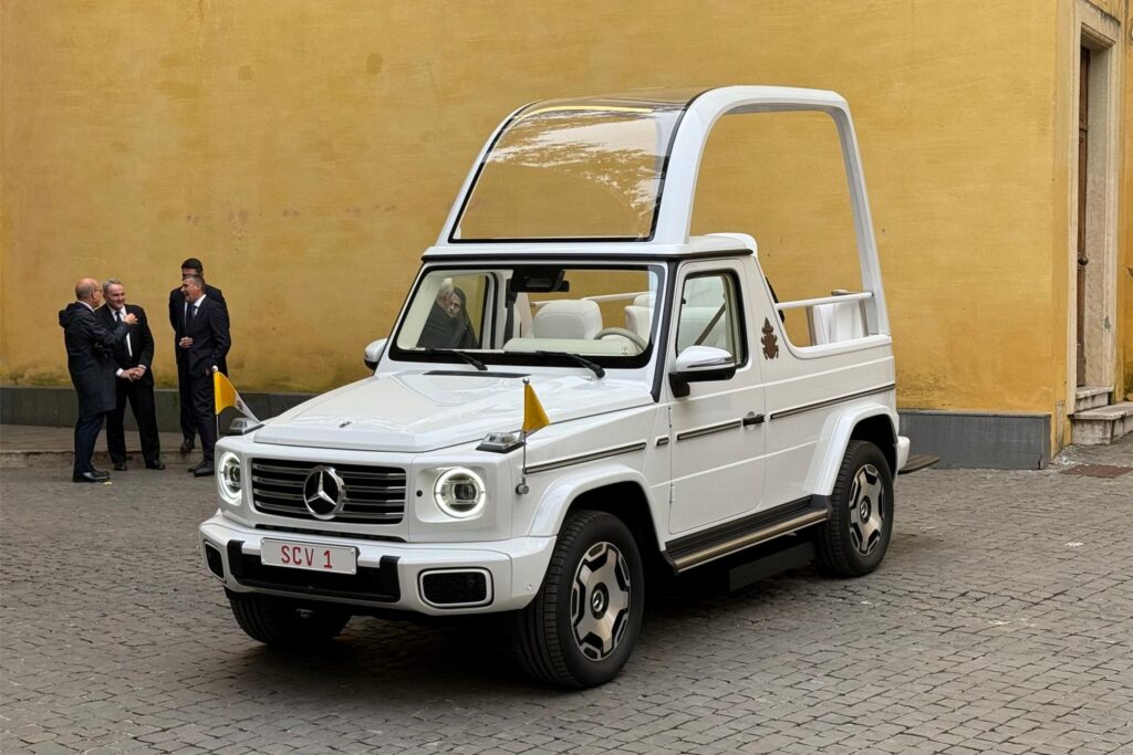 Das weiße Auto des Papstes steht auf einem Platz voll Kopfsteinpflaster, links davon ein paar Männer in Anzügen. Das weiße Auto des Papstes steht auf einem Platz voll Kopfsteinpflaster, links davon ein paar Männer in Anzügen.