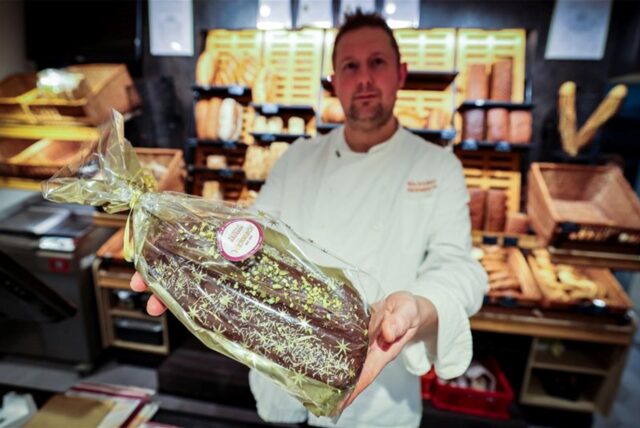 Ein Mann steht in einer Bäckerei und hält einen rechteckigen Stollen in die Kamera.