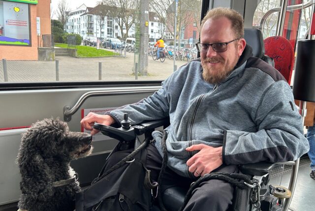 Kai Gungl sitzt im Rollstuhl in einer Stadtbahn mit seinem Assistenzhund Bella (12).