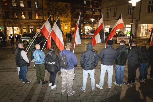 Eine Gruppe junger Erwachsener in Winterkleidung, teils mit Kapuzen, steht auf einem gepflasterten Platz in einem Halbkreis. Sie stehen mit dem Rücken zur Kamera, sechs von ihnen