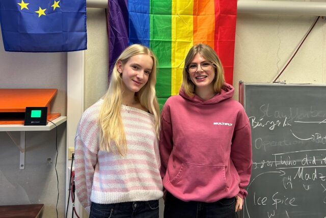Zwei Frauen stehen im Klassenraum im Theodor-Heuss-Gymnasium in Waltrop vor einer Regenbogen-Flagge.