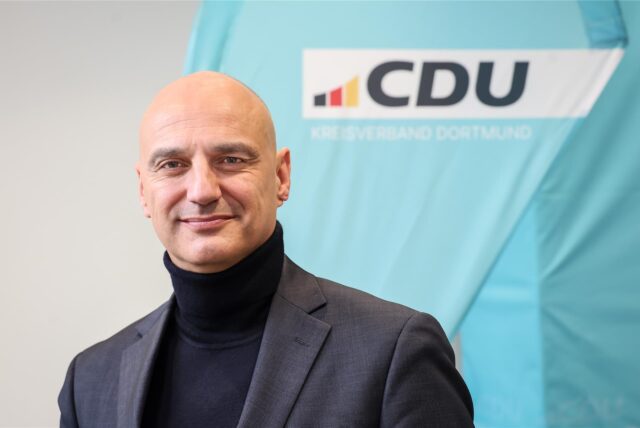 Alexander Kalouti lächelt in die Kamera, vor einem Logo der CDU