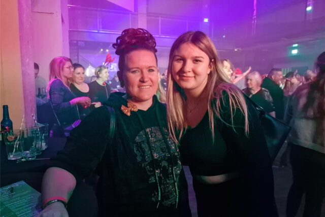 Zwei Frauen stehen in der Maschinenhalle der Zeche Ewald in Herten-Süd auf der Tanzfläche und schauen die Fotografin freundlich lächelnd an. Im Bildhintergrund tanzen viele Männer und Frauen