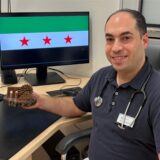 Omar Alubaissi in seiner Arztpraxis vor einer Syrienflagge auf dem Bildschirm und einem kleinem Andenken aus Holz an die Wasserräder seiner Heimatstadt Hama.