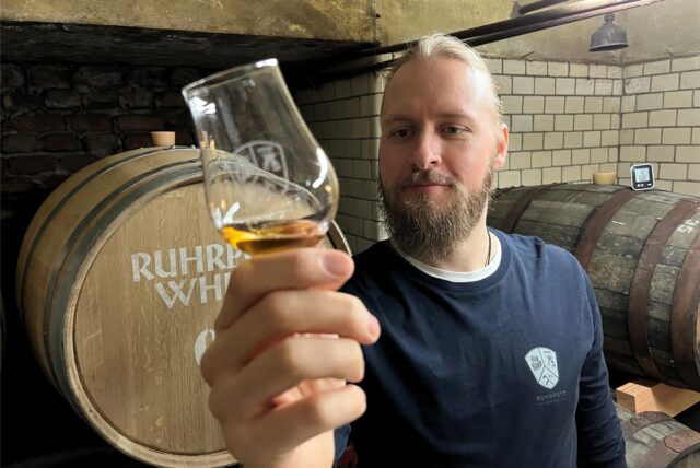 Björn Nessmann blickt auf eine goldene Flüssigkeit in einem kleinen Glas. Dabei steht er vor Fässern  mit dem Aufdruck Ruhrpott Whisky.
