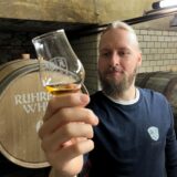 Björn Nessmann blickt auf eine goldene Flüssigkeit in einem kleinen Glas. Dabei steht er vor Fässern  mit dem Aufdruck Ruhrpott Whisky.