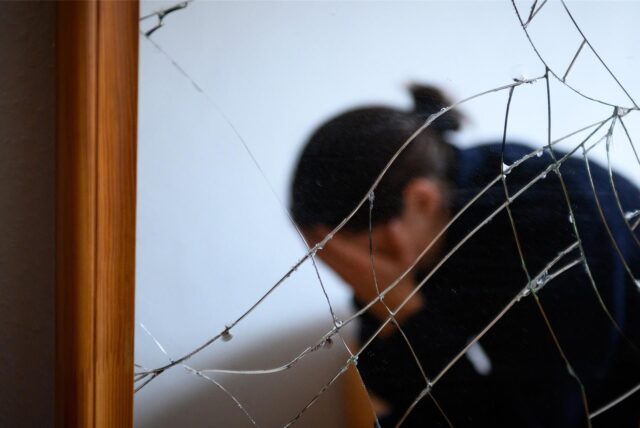 Eine Frau vergräbt ihr Gesicht in ihren Händen. Sie sitzt hinter einer zerstörten Glasscheibe.