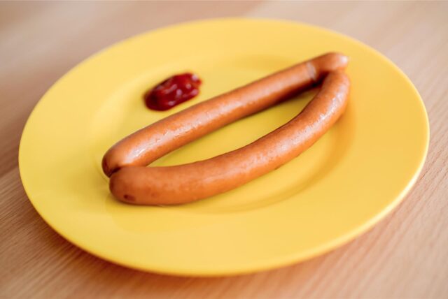 Ein gelber Teller aus Hartplastik mit zwei Würstchen und Ketchup steht auf einem Tisch.