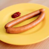 Ein gelber Teller aus Hartplastik mit zwei Würstchen und Ketchup steht auf einem Tisch.