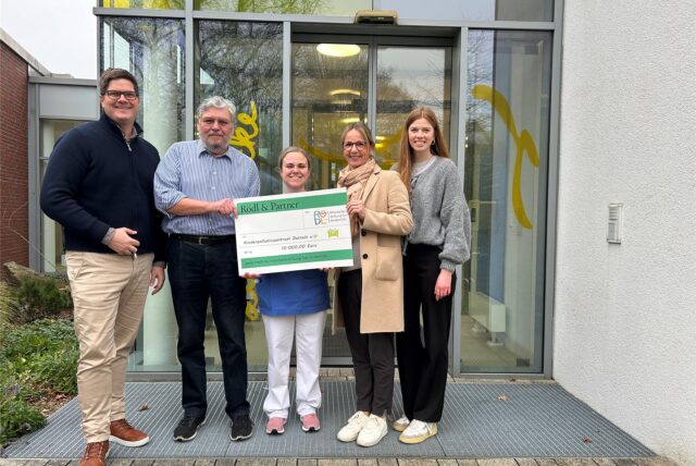 Fünf Personen stehen mit einem großen Spendenscheck vor dem Eingang des Kinderpalliativzentrums.