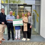 Fünf Personen stehen mit einem großen Spendenscheck vor dem Eingang des Kinderpalliativzentrums.