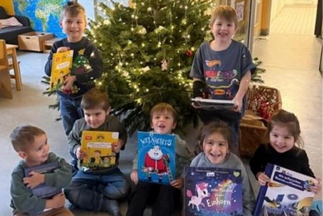 Sieben Kinder stehen und sitzen vor einem Weihnachtsbaum und halten Bücher in der Hand.