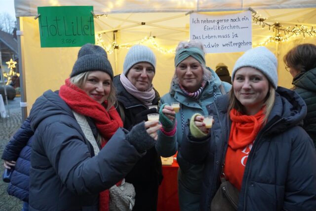 Vier Frauen stehen nebeneinander lächeln in die Kamera und stoßen mit Eierlikörpinnchen an.