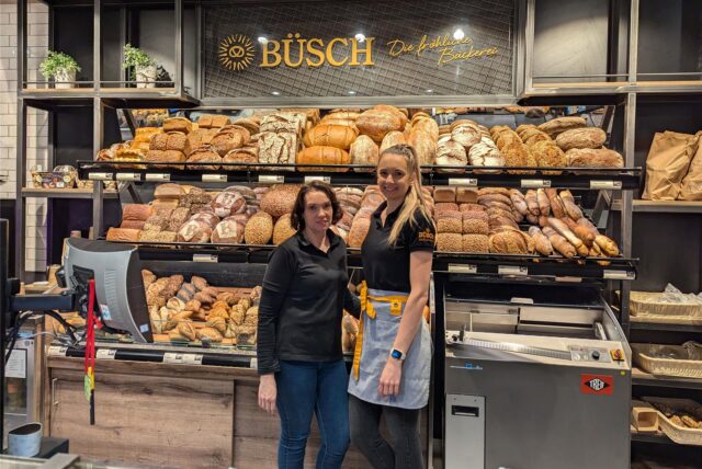 Verkäuferinnen der Bäckerei Büsch stehen in einer Filiale vor der Brot- und Brötchenauslage.