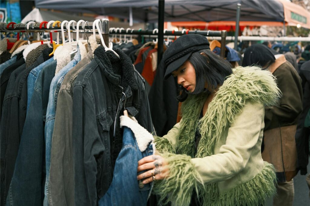 Eine Frau mit einer grünen Jacke und einer schwarzen Kappe durchstöbert einen Stand mit Jacken auf einem Trödelmarkt.