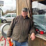 Ein Mann in brauner Fleecejacke lehnt an der Theke einer Verkaufshütte in Waltrop. Vor ihm liegt eine Säge. Hinter ihm ist ein grauer Trichter, um die Weihnachtsbäume einzunetzen.