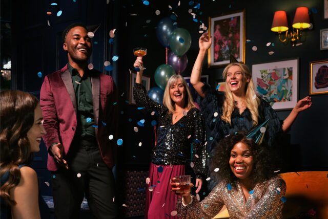 Vier Frauen und ein Mann in festlicher Kleidung und Partyhüten feiern Silvester.