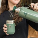 Eine Frau hält eine Thermoskanne und einen Becher von Stanley in den Händen.