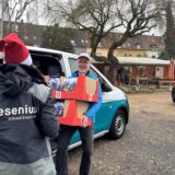Im Vordergrund steht eine Person mit Weihnachtsmütze am linken Bildrand. Auf der Jacke der Person steht Fresenius Umwelttechnik. Neben der Person lädt ein Mann in hellblauer Jacke zwei Pappkartons mit Lebensmitteln aus einem Van. Der Mann lacht in die Kamera. Im Hintergrund sind Gartenmöbel und Wohnhäuser zu erkennen, sowie ein geparkter Van der Hertener Tafel.