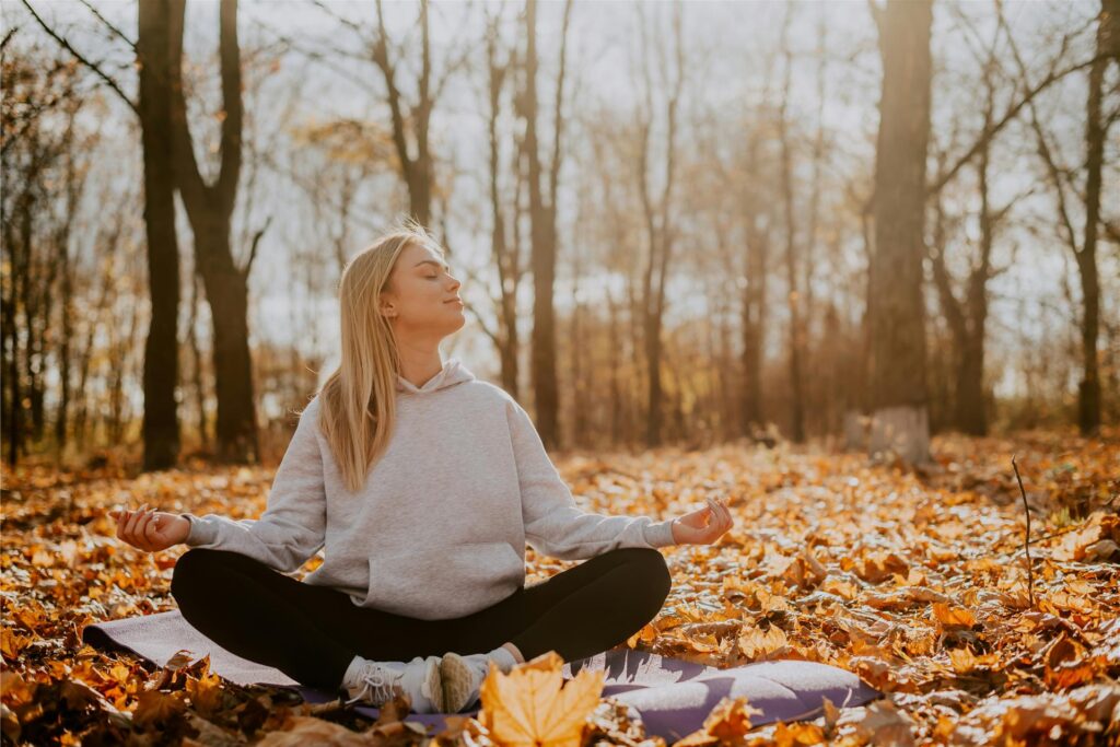 Eine Frau sitzt im Herbst im Wald auf einer Yoga-Matte und meditiert. Mit geschlossenen Augen hält sie ihr Gesicht in die Sonne. Eine Frau sitzt im Herbst im Wald auf einer Yoga-Matte und meditiert. Mit geschlossenen Augen hält sie ihr Gesicht in die Sonne.