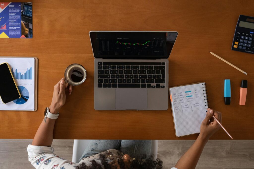 Eine Frau sitzt mit ihrem Laptop, ihrem Notizbuch und einem Kaffee an ihrem Schreibtisch und verwaltet ihre Finanzen.