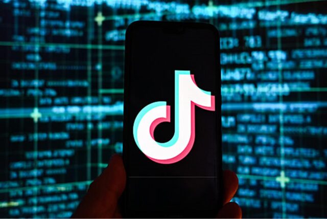 Das Logo der Video-App TikTok ist auf einem Smartphone vor einem Bildschirm mit Börsenkursen im Hintergrund zu sehen.