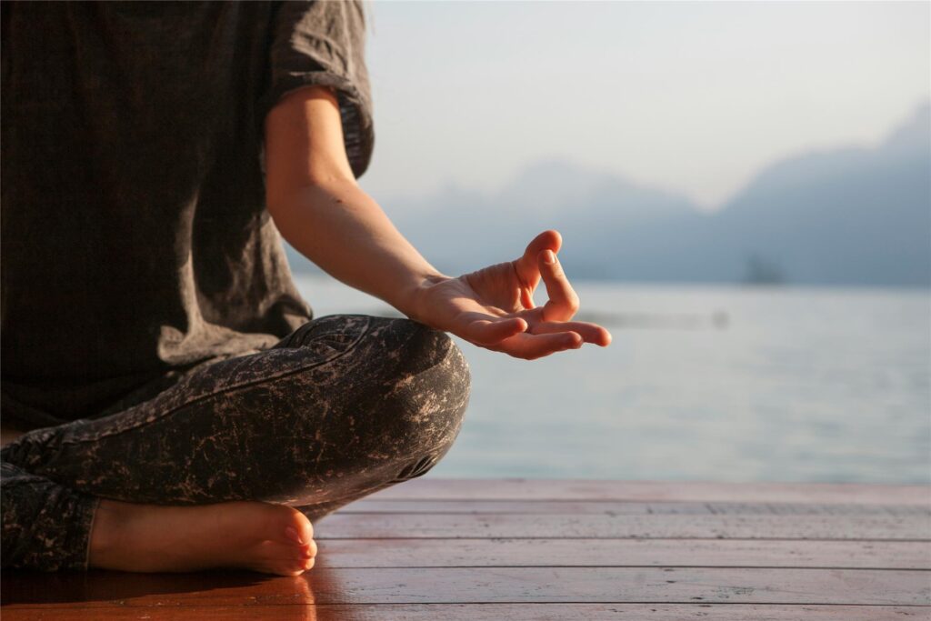 Eine Frau sitzt auf einem Holzsteg an einem Bergsee und macht Yoga.