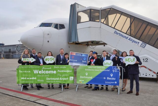 Auf dem Rollfeld des Dortmunder Flughafens posieren vor einem Flugzeug Mitarbeiter aus Dortmund und Rumänien. Vor ihnen stehen Banner mit der Aufschrift: Willkommen, Dortmund, Bacău, jetzt mit Dan Air.
