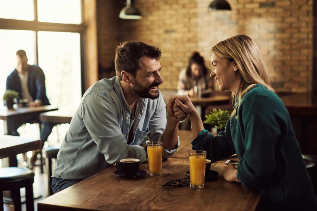 Ein Mann und eine Frau sitzen sich bei einem Date gegenüber, lachen und halten sich an der Hand. Zwischen ihnen auf dem Tisch stehen zwei Kaffeetassen und zwei Gläser mit Orangensaft.